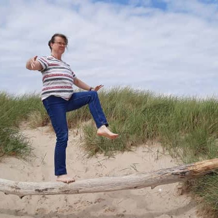 Ulrike Stähr balanciert barfuß auf einem Baumstamm vor einer Graslandschaft.